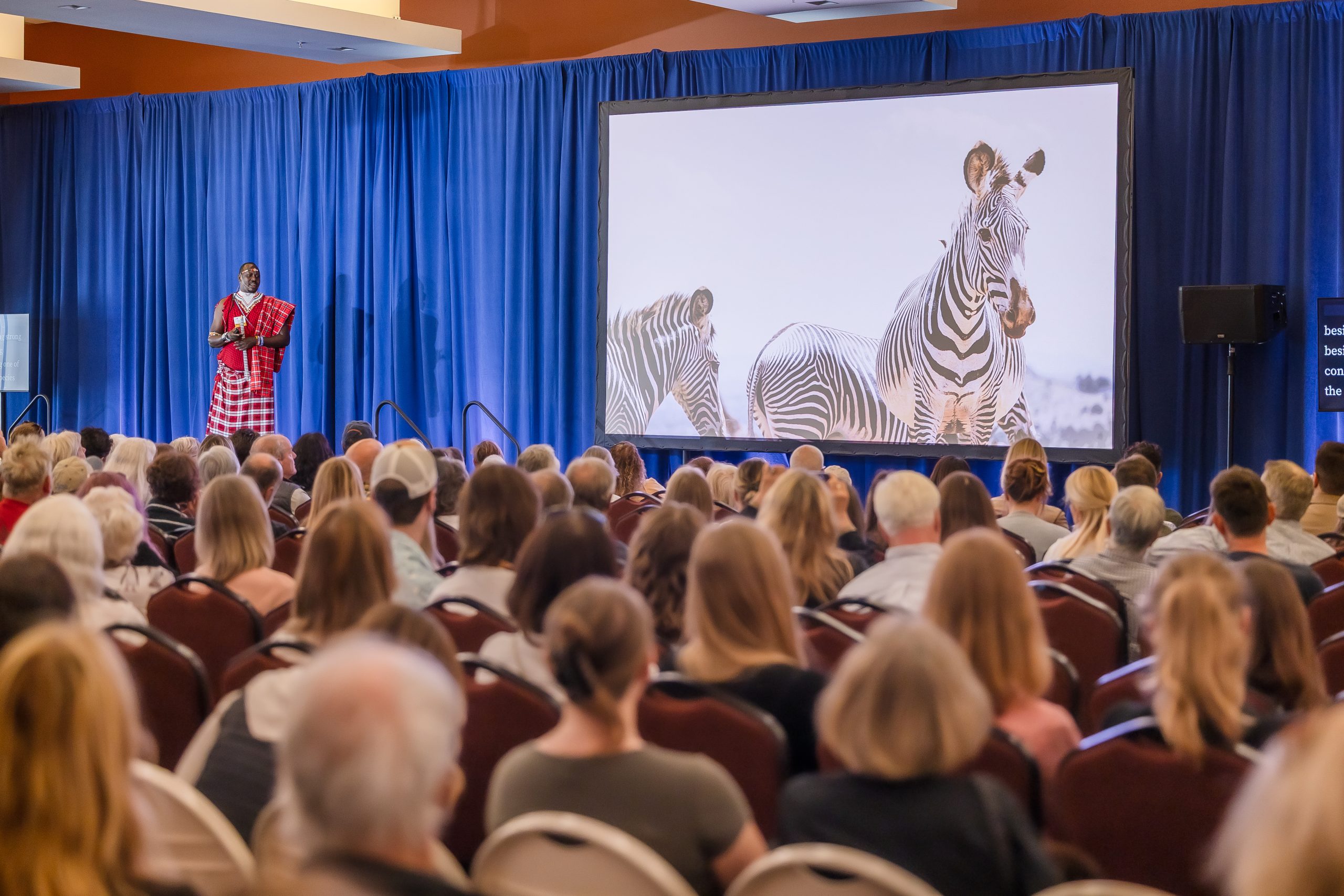 Peter Lalampaa, Director of Programs at Grevy’s Zebra Trust, presenting on stage at WCN’s 2025 Expo at the Mission Bay Conference Center in San Francisco, with an image of zebras on the screen and a full house of attendees in the audience.